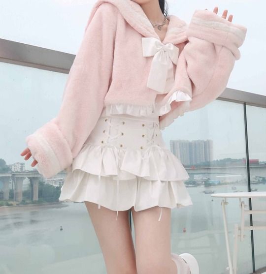 a young woman poses on a balcony, showcasing a stylish outfit. she wears a cropped pink fuzzy jacket with a large white bow and a white skirt with ruffles. her outfit exudes a cute and fashionable aesthetic, often seen in lolita or kawaii fashion. the background reveals a glimpse of a city under a cloudy sky.