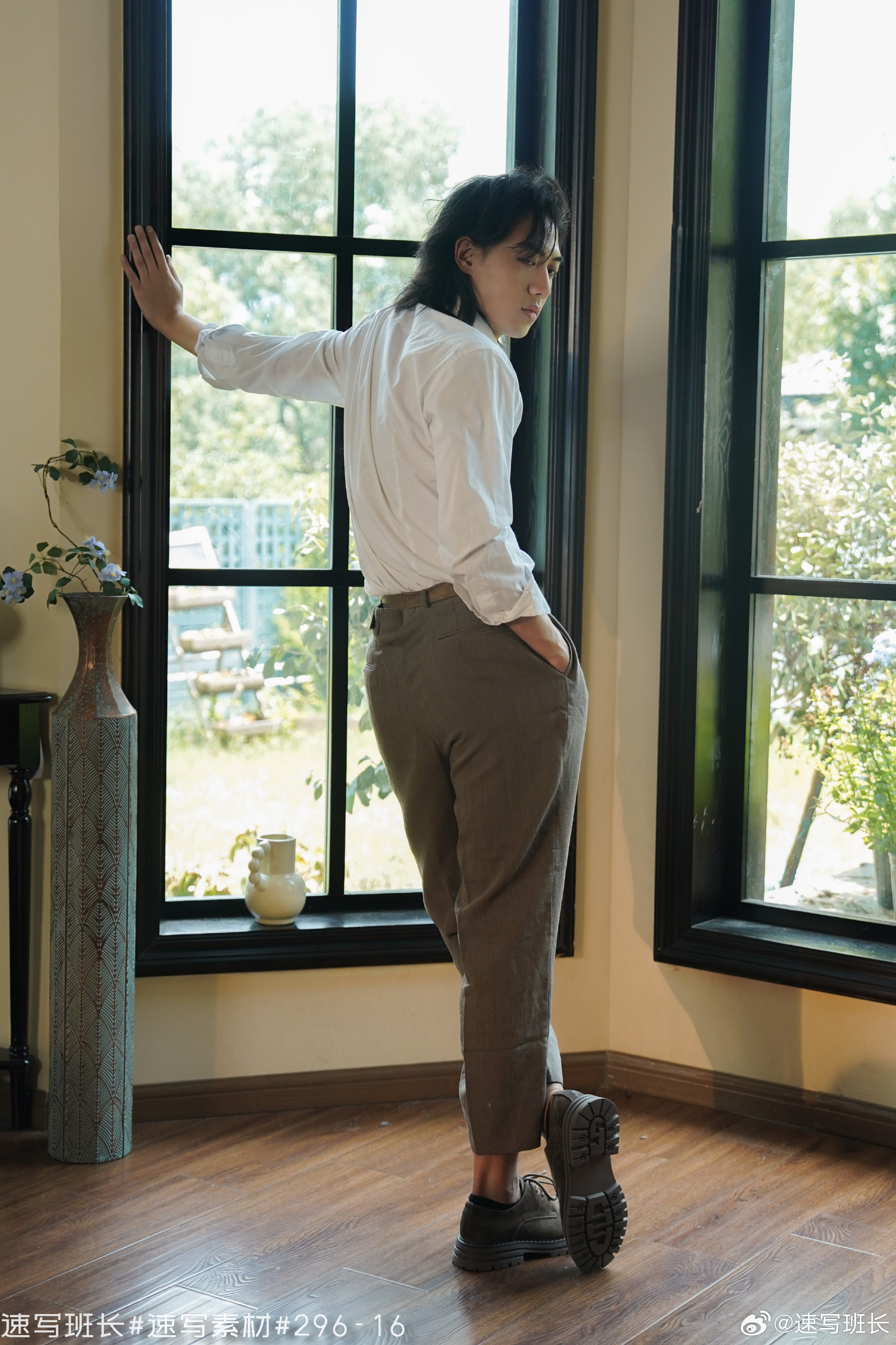 a man with long black hair is standing in a room, looking out of a large bay window. he is wearing a white shirt, brown pants, and black shoes. the room has wooden floors and white walls. there is a vase of flowers on a table in the foreground.