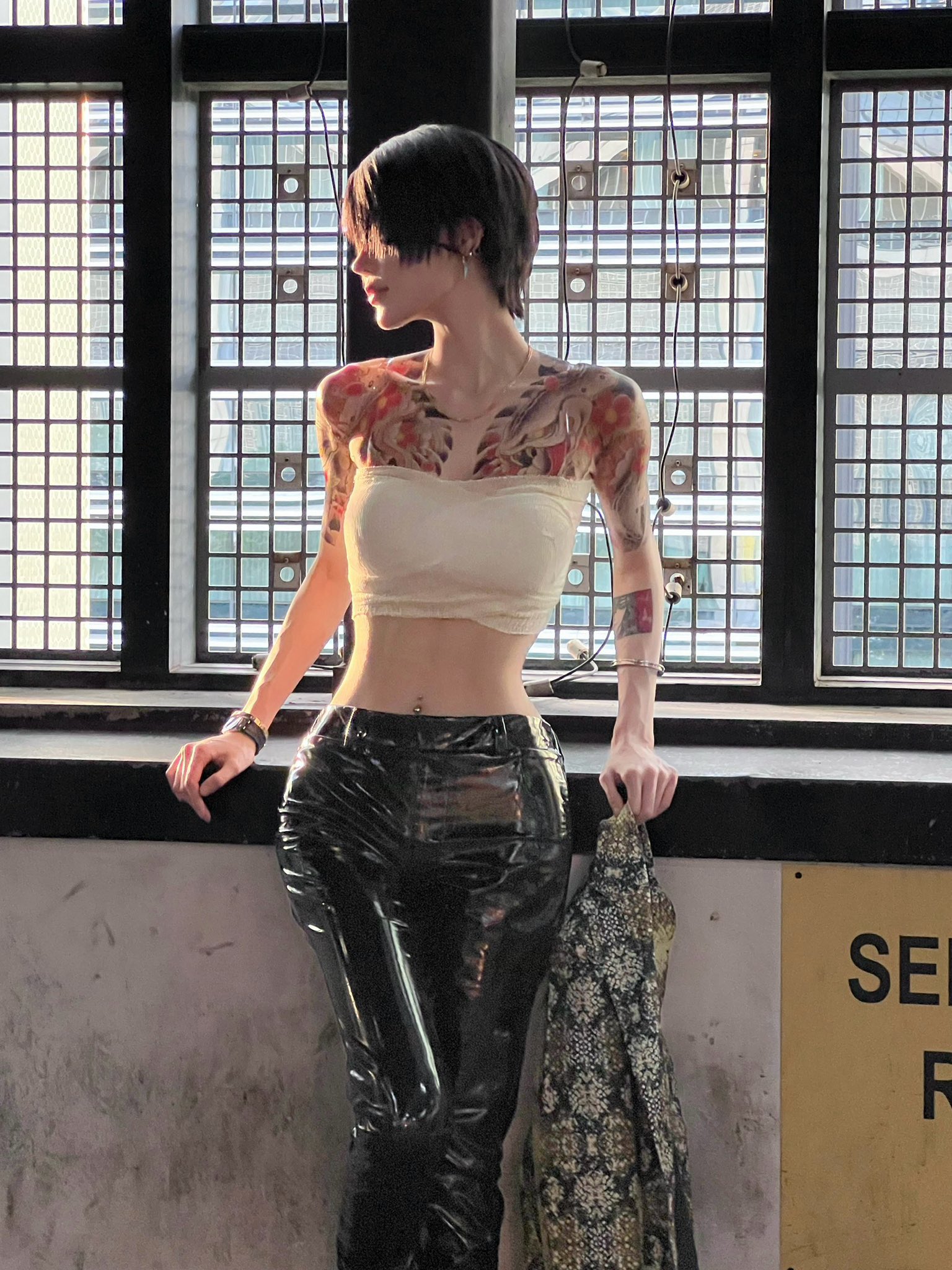 a stylish young woman with tattoos on her arms and chest is standing by a large window in a modern building. she is wearing black vinyl pants, a creamcolored crop top, and holding a patterned jacket.  the image highlights her slim figure and fashionable outfit. the setting is urban and contemporary.