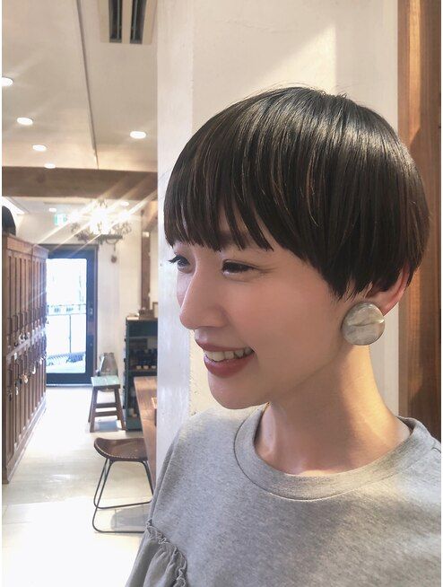 a woman with short brown hair is smiling while looking to the left. she is wearing a grey sweater and large, grey earrings. she is in an indoor space, possibly a salon.