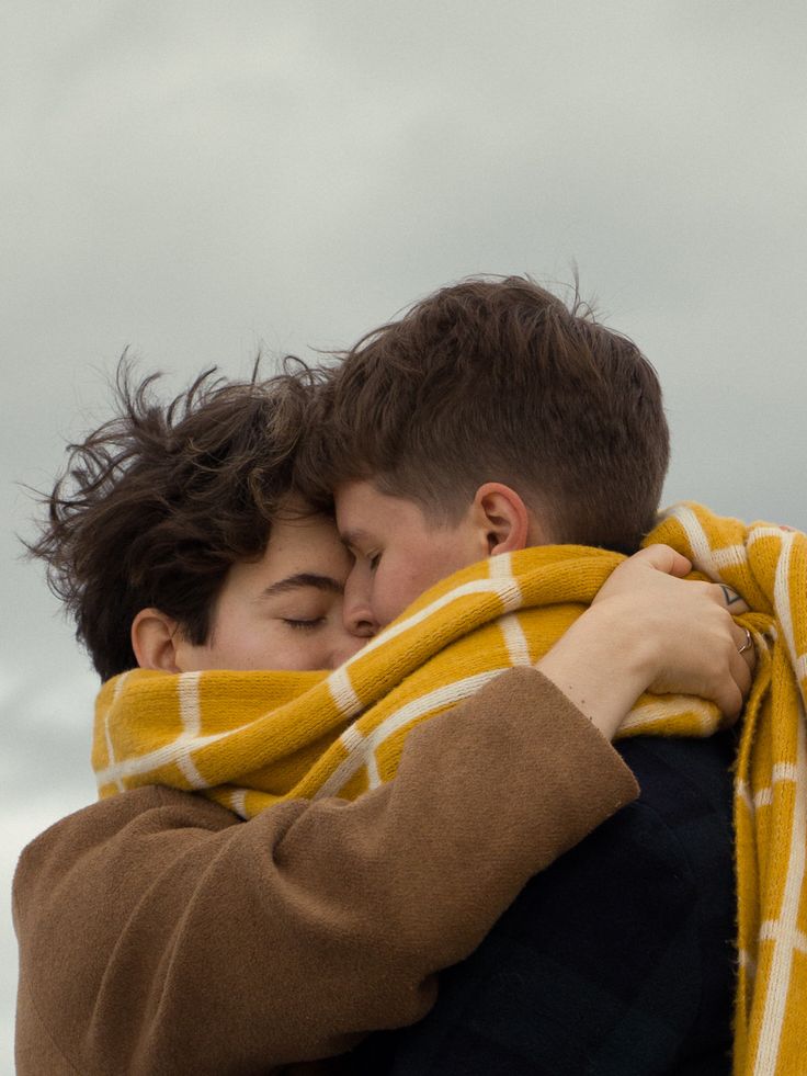 two men are embraced in a tender hug. they are both wearing brown jackets and a yellow and white scarf. their eyes are closed and they appear to be enjoying each other's company.  the men are outdoors on a cloudy day.