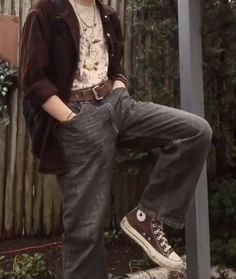 a person wearing a brown jacket, brown pants, and brown converse shoes. they are standing in front of a wooden fence and are looking down at the ground. the image has a vintage aesthetic.