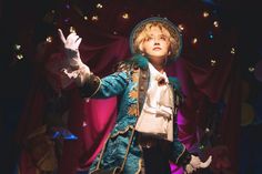 a man in an elaborate stage costume stands with one hand raised against a backdrop of red curtains and twinkling lights. he appears to be a performer, possibly a magician, in the middle of a performance.