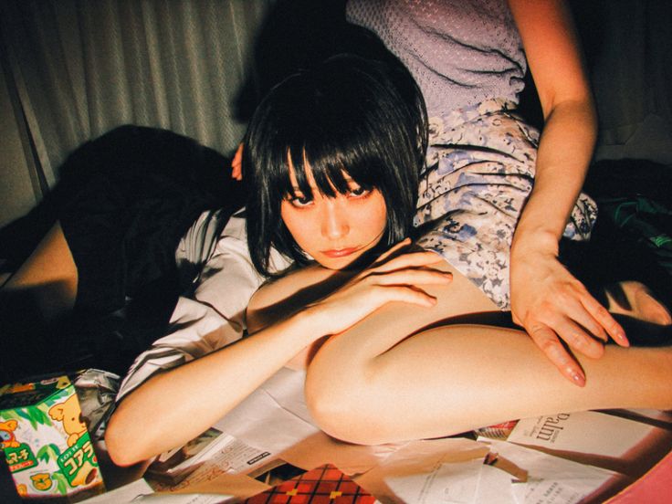 two young japanese women in a bed in a bedroom.  the woman in the back is wearing a white shirt and a dark skirt. she is sitting up, looking down with her hands on her legs. the woman in the front is wearing a blue shirt and a light colored skirt with white flowers. her legs are spread out in front of her and she is resting her head on her hands. the image is shot from a low angle, and there is a soft light illuminating the subjects.  it is a casual and intimate portrait of two young women.