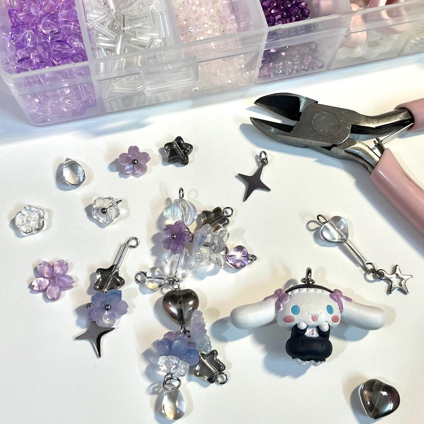 a highangle shot showcases a variety of craft supplies spread out on a white surface. at the top is a clear plastic container divided into compartments, each filled with different types of beads in shades of purple, clear, and silver. various charms are scattered around, including flower charms in purple and white, star charms in silver, and a cinnamoroll charm, which is a white doglike character with purple bows. there are also heartshaped charms in silver. crafting pliers with pink handles are positioned to the right of the beads. the image suggests a jewelrymaking project or hobby in progress.
