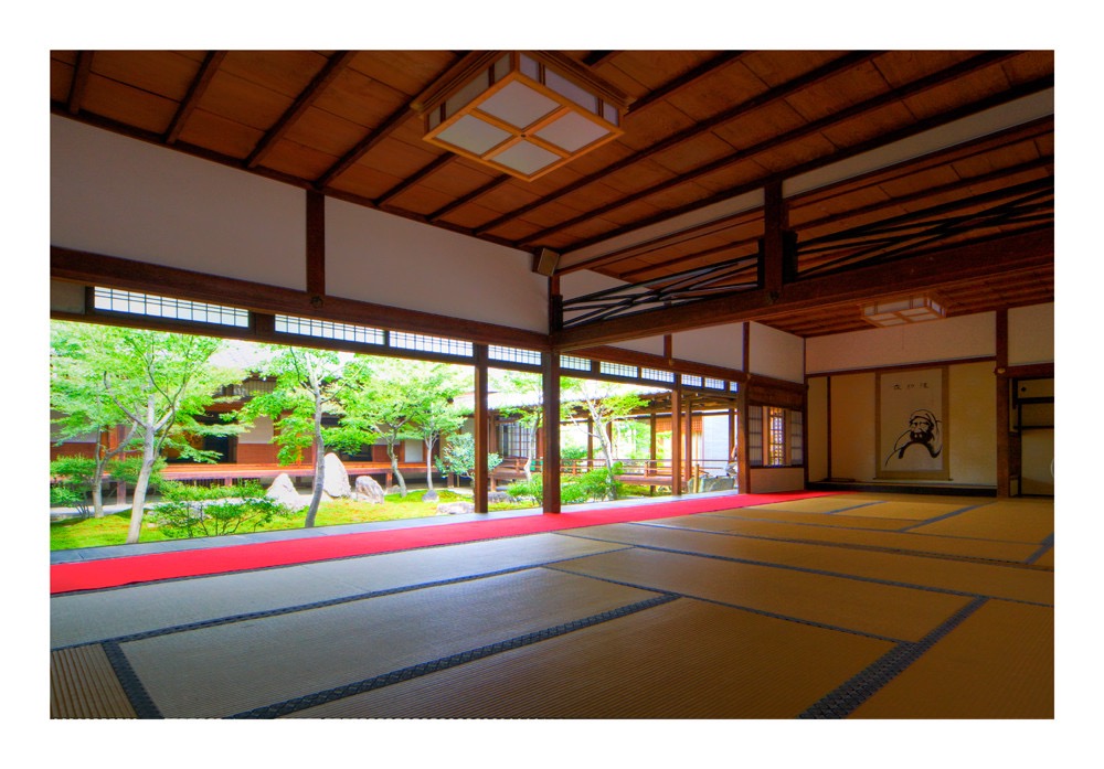 a view from the inside of a traditional japanese home, looking out onto a zen garden. the room features tatami mats on the floor, wooden beams on the ceiling, and large windows that frame the green and lush garden. a red rug lies in front of the windows, adding a pop of color to the serene scene. the natural light pours in through the windows, illuminating the space.
