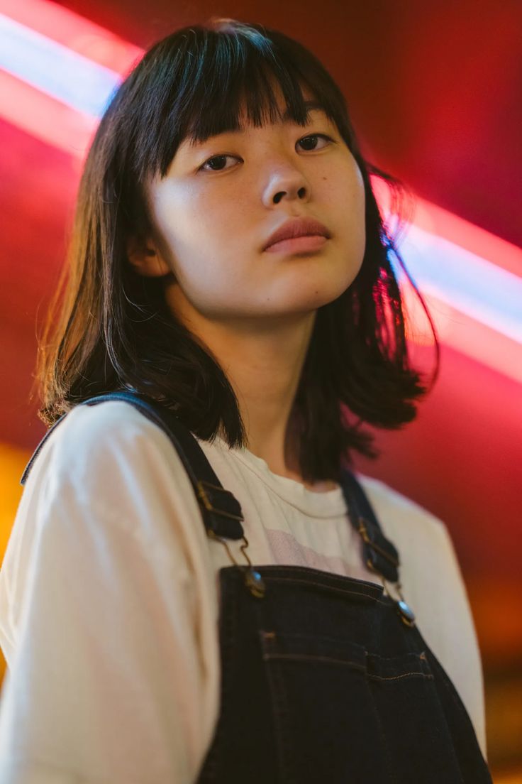 a medium shot portrait of a young asian woman with dark hair and bangs. she is wearing a white tshirt and dark denim overalls. the background features a bright red and blue neon light. she is looking directly at the camera.