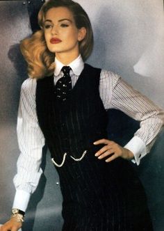 a woman in a business suit with a vest and tie. she is standing in a studio and looking at the camera with a confident expression. her hairstyle is a classic blonde bob, and her makeup is subtle yet chic. she is wearing a white shirt with black stripes under her suit vest, and her vest has black stripes on it. she is also wearing a tie that is a dark shade of red and has a pattern that is difficult to make out. the woman is wearing a silver bracelet on her left wrist and a ring on her right hand. the background is a dark grey color, and the image is focused on the woman's face and upper body. her outfit is very professional and sophisticated, and it is perfect for a business meeting or other formal event.