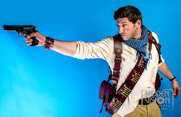 a man dressed as nathan drake from the uncharted video game series, holding a gun and pointing, standing in front of a blue background.