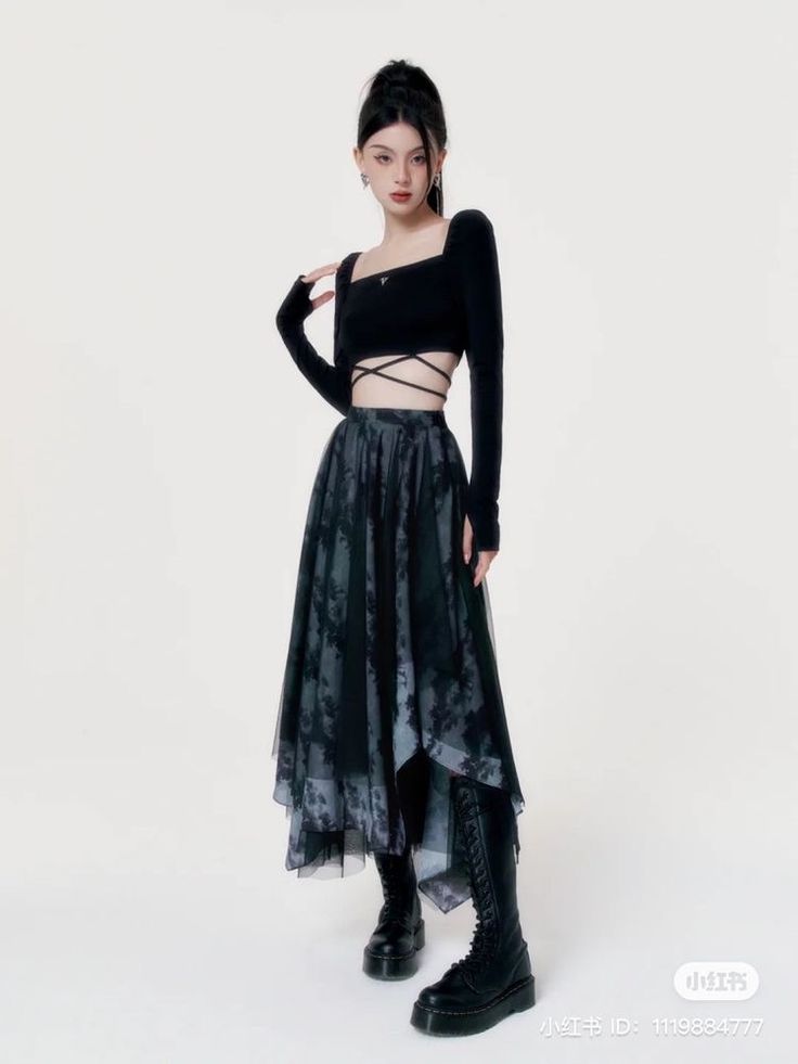 a full shot of a young woman posing in a black longsleeved crop top and a darkcolored, asymmetrical tulle skirt. the skirt has a pattern that resembles tiedye. she is wearing black laceup boots and has her hair pulled back. the background is plain white.