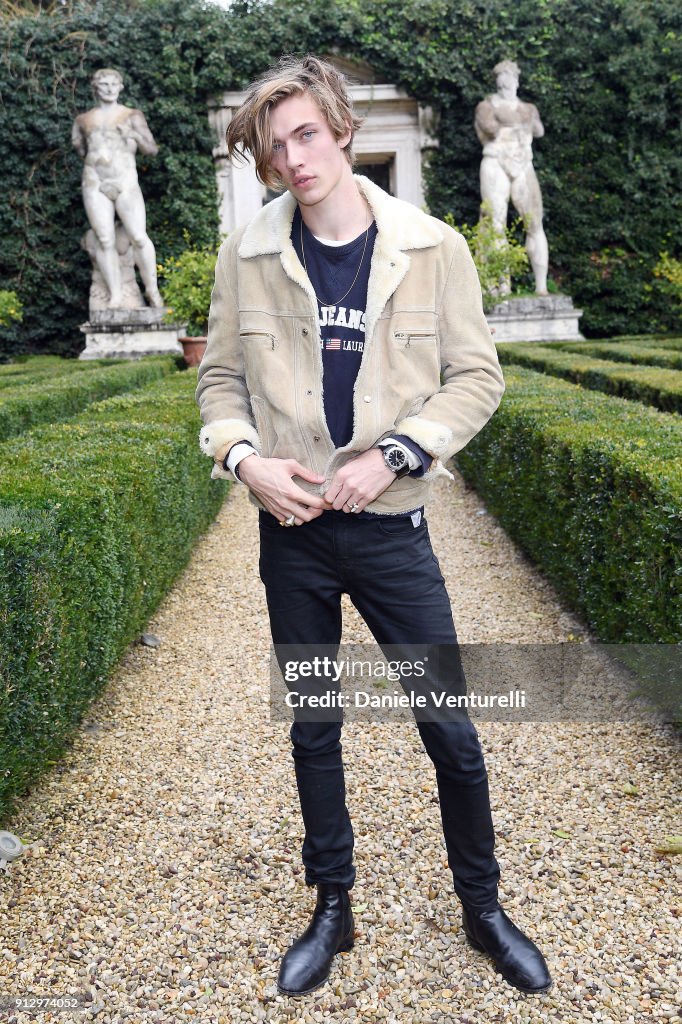 a full shot of a young man standing on a gravel path in a garden. he is wearing a light beige jacket with a white collar, a dark tshirt, black jeans, and black boots. he is also wearing a wristwatch. behind him is a hedge, a statue, and a building. the image is a getty images photograph by daniele venturelli.