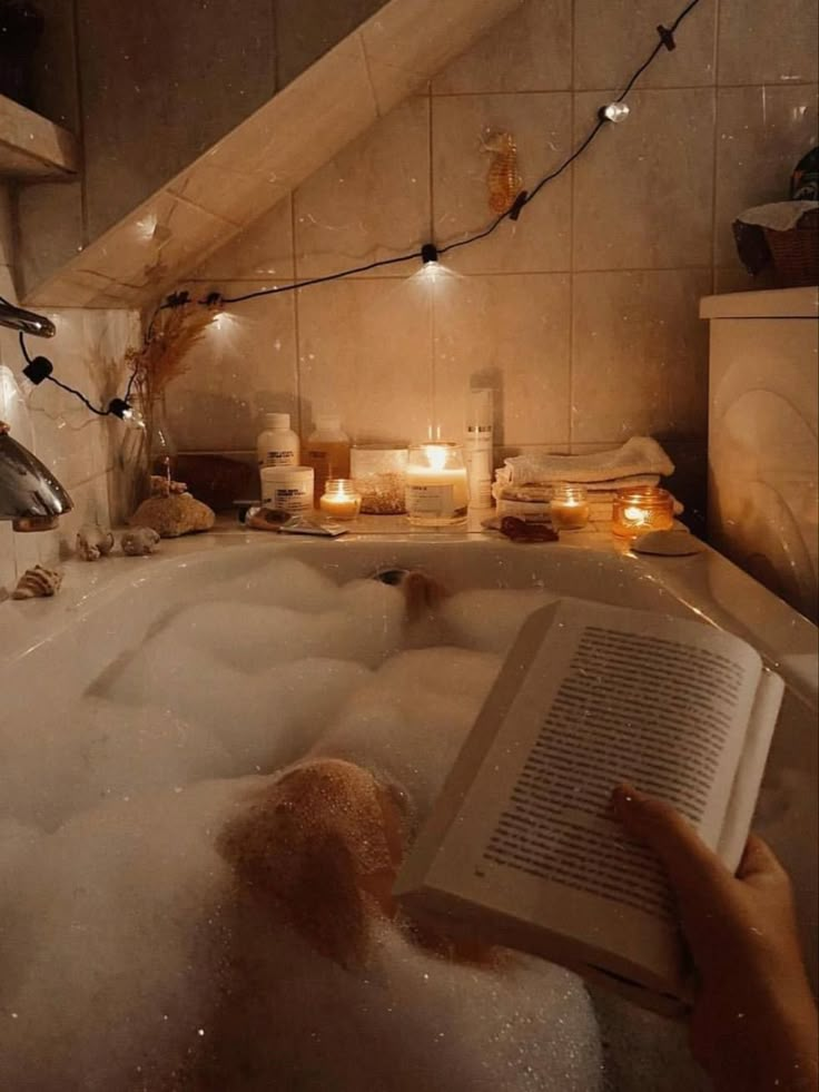 a person is relaxing in a bubble bath, reading a book. the bathroom is decorated with candles and string lights to create a cozy and relaxing atmosphere. there are beauty products on the side of the tub.