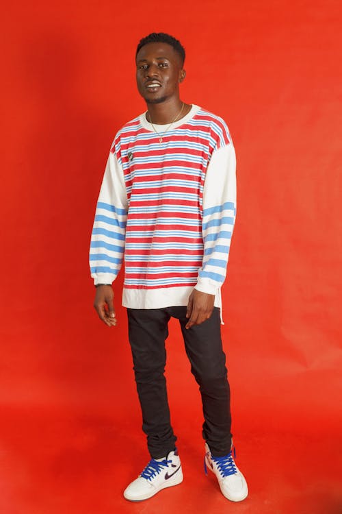 a fullbody shot of a man standing against a red background. he is wearing a longsleeved shirt with red, white, and blue horizontal stripes, black jeans, and white sneakers with blue laces. he is also wearing a necklace.