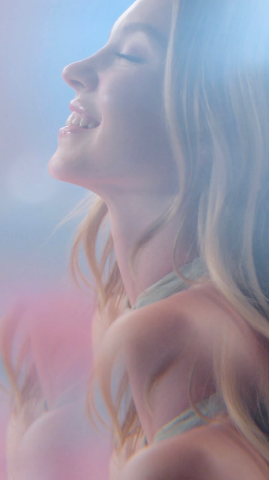 a closeup shot of a woman with long blonde hair, her eyes closed in a serene and peaceful expression.  she is smiling gently, and her skin appears soft and glowing. the image has a dreamy, ethereal quality due to the soft focus and pastel pink and blue color palette, creating a romantic and sensual atmosphere.  the lighting is soft and diffused, enhancing the overall ethereal effect.  it's likely an image for a beauty or fashion advertisement.