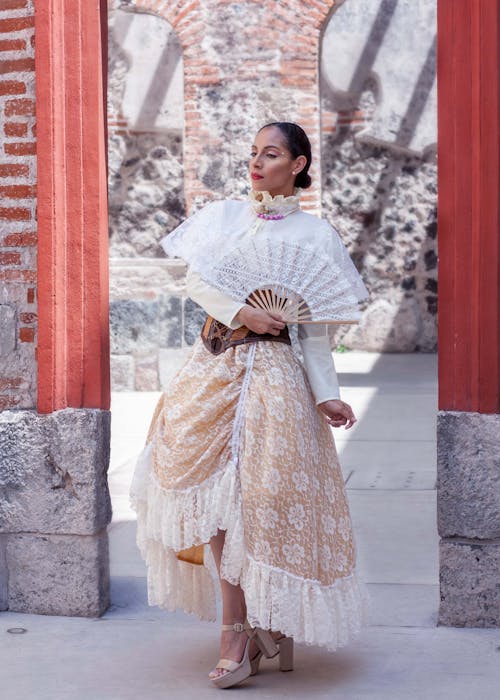 a woman in a beautiful vintagestyle lace dress stands in a doorway of an old brick building. she is holding a lace fan and wears beige high heels.  the dress is a cream and beige color with layers of lace, and is styled in a highlow hemline. the setting appears to be a historical location, possibly in mexico.  she's posed elegantly, and the overall image has a romantic and nostalgic feeling.