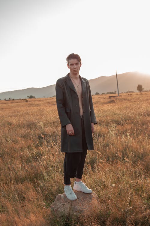 a young man stands in a tall grass field at sunset, wearing a long dark coat and white sneakers. he has a serious expression and is looking directly at the camera. the setting sun casts a warm glow on the scene.  he appears to be standing on a small rock.  the overall atmosphere is serene and contemplative.