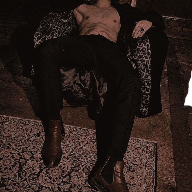 a man wearing a black suit, sitting on a leopard print couch with a ring on his finger. he is shirtless and has abs, and the lighting in the room is dark.  he is wearing brown boots.
