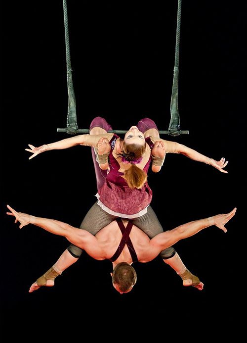two acrobats are performing a trapeze act. the female acrobat is upside down, her legs wrapped around the male acrobat's waist. the male acrobat is holding on to the trapeze bars with his hands and feet, while the female acrobat is holding on to the trapeze bars with her legs. they are both smiling and looking up at the audience. the background is a black curtain.  the performers are wearing red and black costumes.
