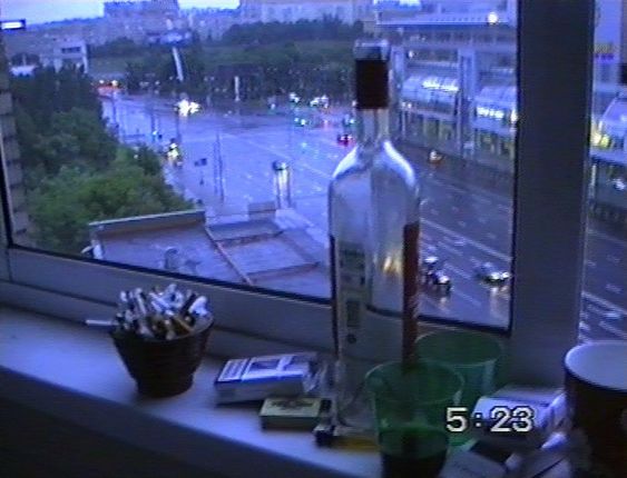 the image is a grainy picture taken from a window looking out onto a city street at night.  it is raining and the streetlights reflect off the wet pavement.  on the windowsill are an empty wine bottle, a pack of cigarettes, a lighter, two green glasses, and a brown coffee mug.  the overall tone of the image is blue and gray.
