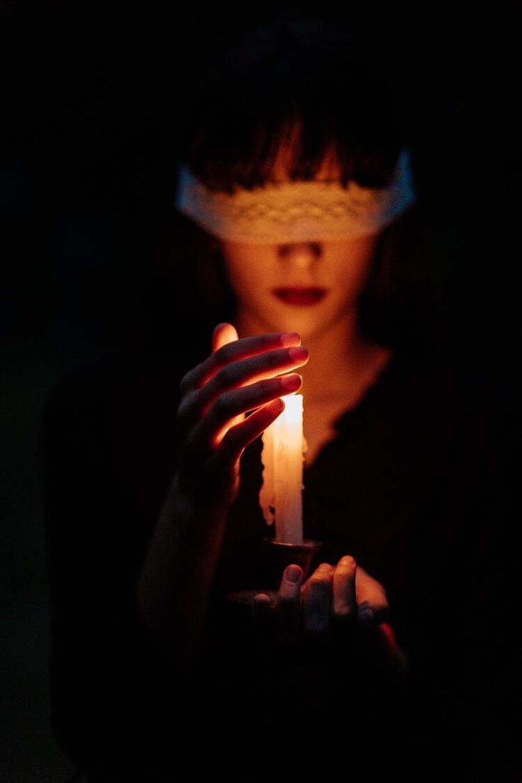 a woman in a dark room holds a lit candle in front of her. she is wearing a blindfold over her eyes. the light from the candle illuminates her face and hands. the room is dark and mysterious. the woman looks scared and alone.