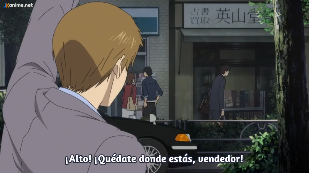 the image is a screenshot from an anime, likely depicting a tense scene. a man in a grey suit with brown hair raises his arm in a commanding gesture, shouting "stop! stay where you are, salesman!" he is likely addressing another man in a suit who is walking away from a storefront labeled "eizando." a black car is parked on the street behind the shouting man. the overall color palette is muted and greyish, contributing to the serious mood.