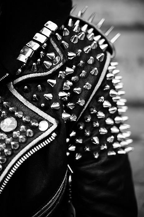 a closeup black and white shot of a black leather jacket heavily studded with metal spikes. the focus is on the shoulder area, showcasing the intricate arrangement of the studs and the texture of the leather. the image evokes a punk rock or rock and roll aesthetic. the zipper and seams of the jacket are also visible.