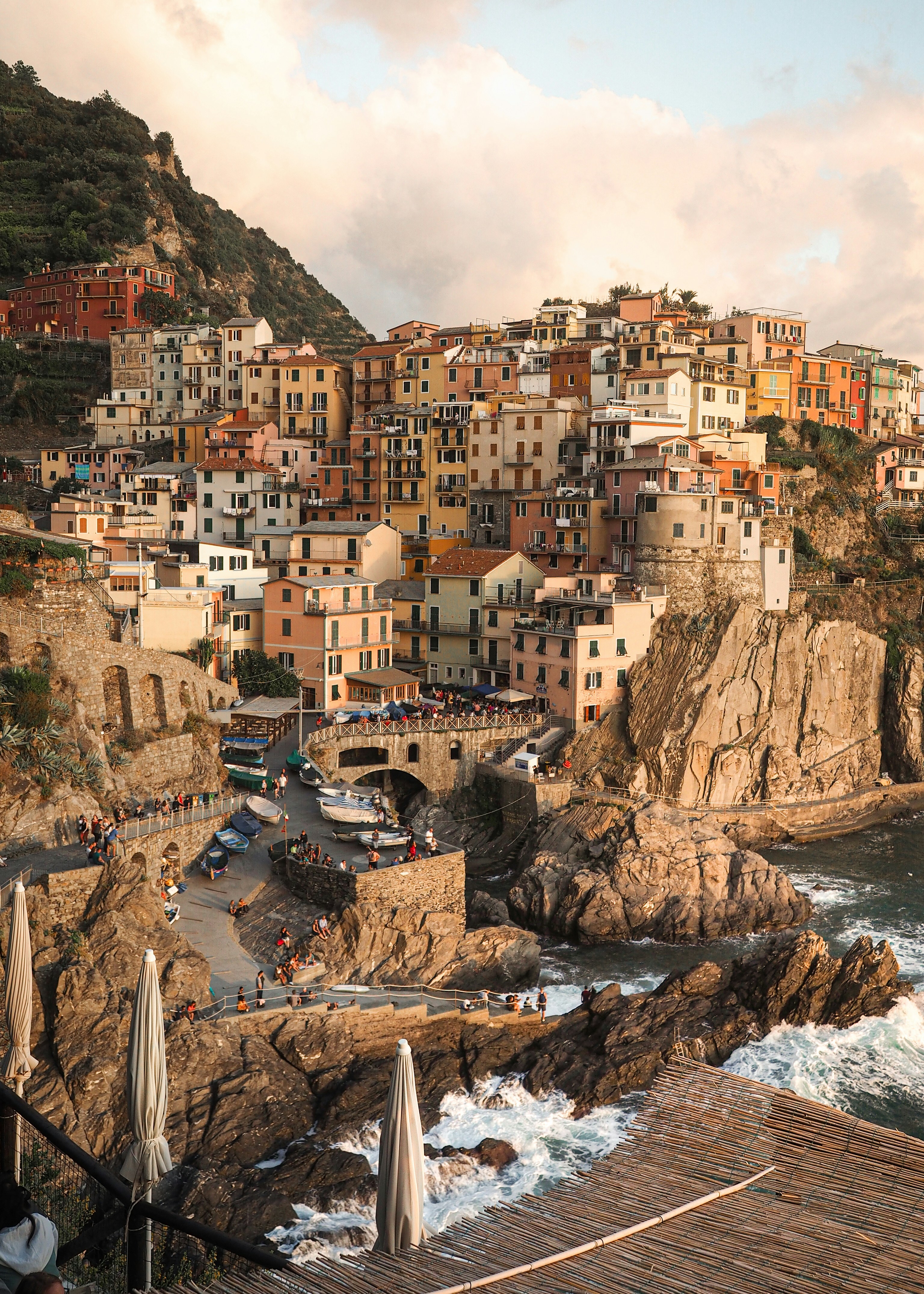 the image shows a picturesque coastal town nestled on the cliffs by the ocean. the town features buildings in various colors stacked upon each other, creating a charming and chaotic landscape. a crowd of tourists gathers on the lower levels, enjoying the sunset over the sea. the lighting is warm and inviting, capturing the beauty of the italian riviera.