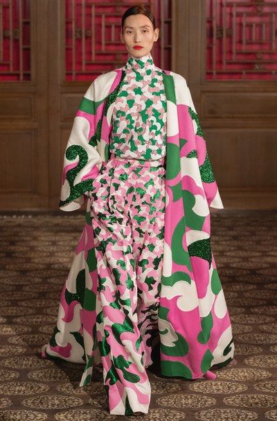 full shot of a model showcasing a striking haute couture ensemble. the outfit comprises a long, flowing coat and matching widelegged trousers, both adorned with a vibrant pink, green, and white floral pattern. the pattern seems to be embroidered or embellished with intricate detailing. the coat features a relaxed fit with dramatic sleeves, while the trousers possess a pleated, flowing silhouette.  the model's sophisticated updo and neutral makeup accentuate the boldness of the clothing. the backdrop is a classic, opulent interior with rich wood paneling and a patterned carpet, suggesting a highfashion runway setting. the overall aesthetic is contemporary yet subtly evocative of vintage styles.