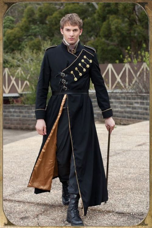 a man stands outside wearing a long black coat with gold trim and buttons, black pants and boots, and holding a walking stick. he appears to be in a regency era inspired outfit. the background features greenery and a brick wall.