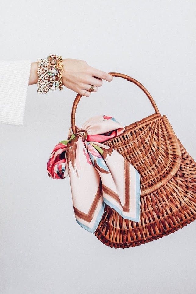 the image shows a woman's hand, adorned with several bracelets, holding a light brown wicker handbag. the bag has a top handle and a slightly curved shape. a pastelcolored floral silk scarf is tied around the handle of the bag. the background is a plain, lightcolored wall. the overall aesthetic is stylish and summery.