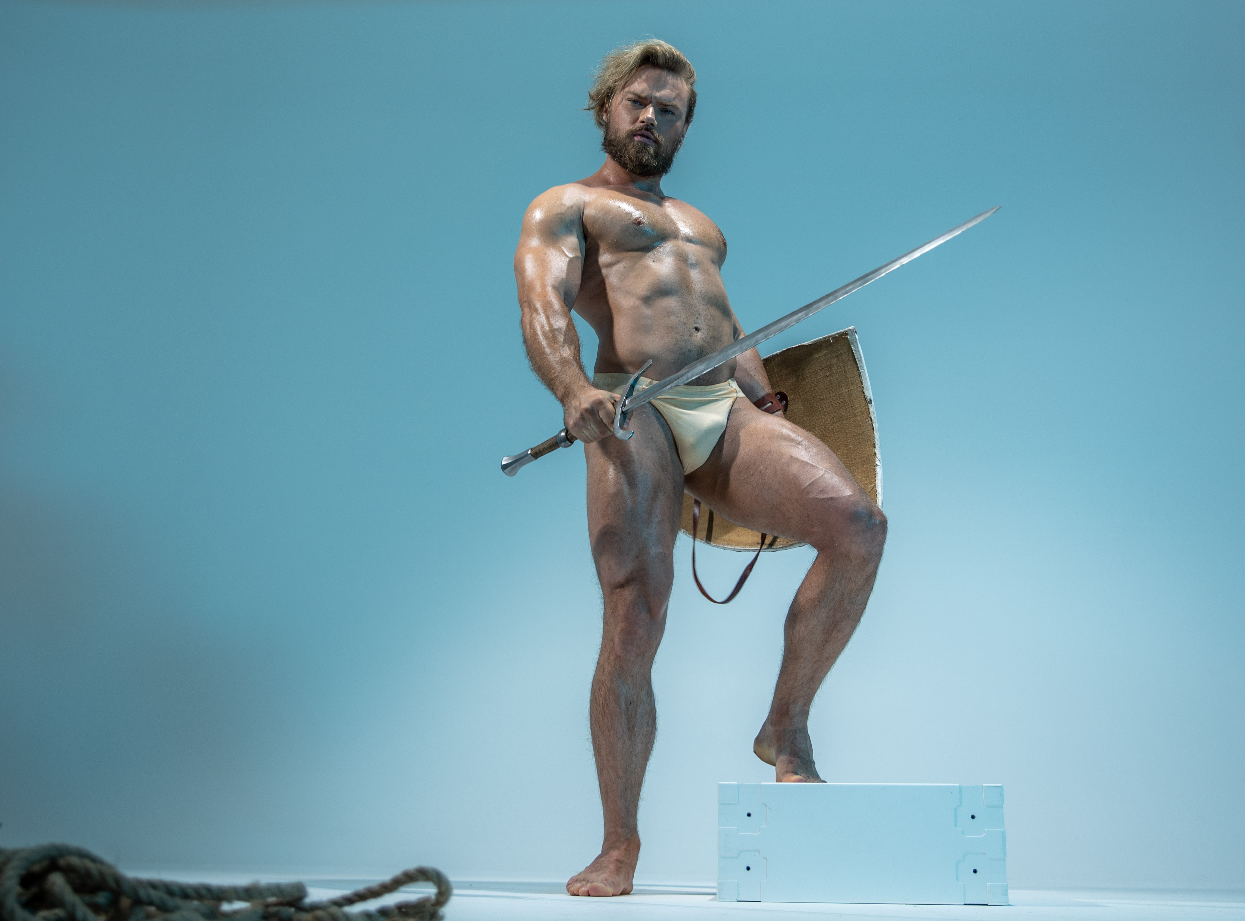 a muscular man with long blond hair stands on a white platform in a studio setting. he adopts a warriorlike pose, holding a sword and shield. he wears only a white loincloth, showcasing his physique. the background is a light blue, and the lighting is soft and even.