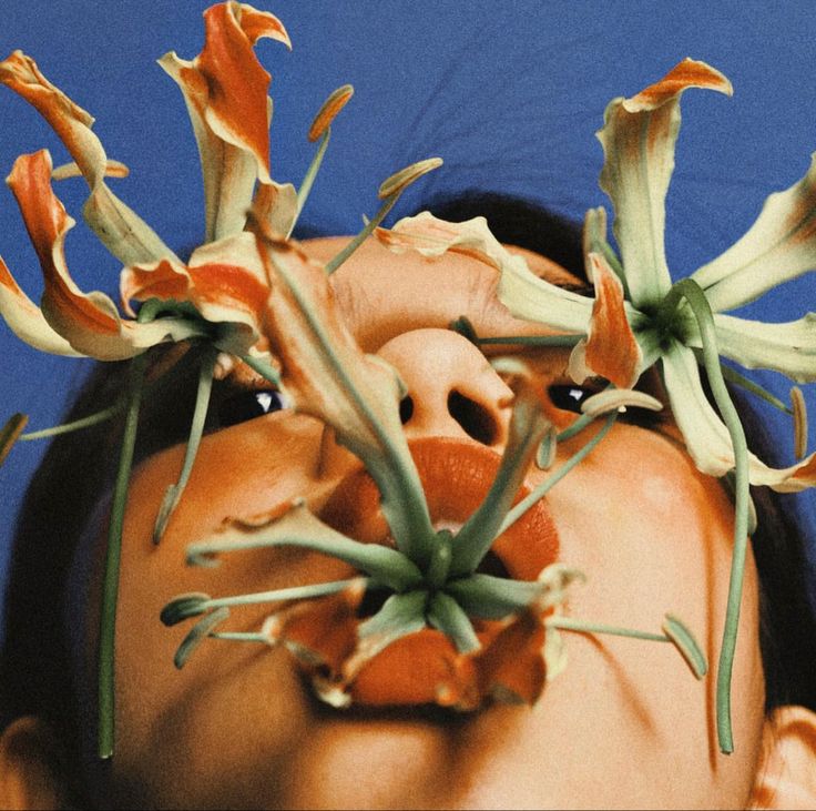 the image is a closeup, lowangle shot of a woman's face with several gloriosa superba lilies (also known as flame lilies or fire lilies) positioned around her mouth and eyes. the lilies' bright orange and red petals contrast sharply against her skin and the green stems create a dramatic visual effect. the woman's expression is intense, her gaze directed upwards. the overall style is artistic and surreal, with a focus on the textures and vibrant colors. the makeup is noticeable, particularly the orange lipstick, which complements the flowers. the image seems professionally shot, possibly for a fashion or beauty publication.