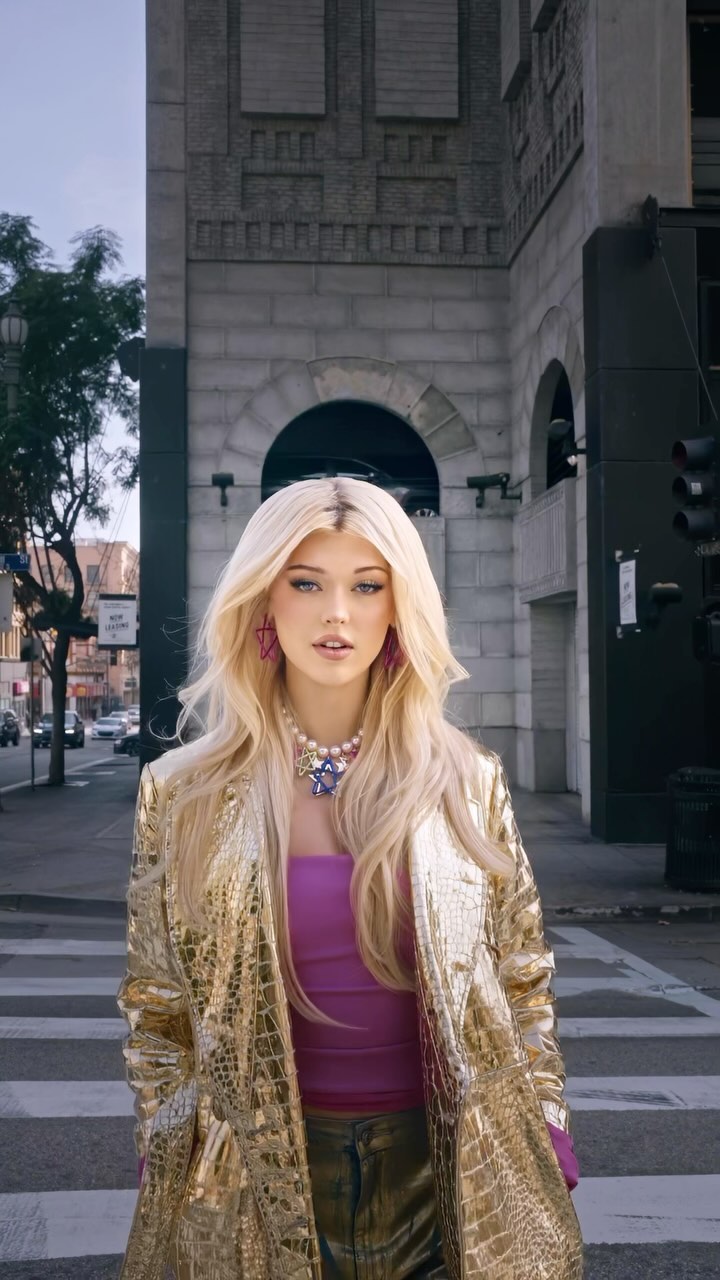 a woman with blonde hair is standing on a zebra crossing in a city street. she is wearing a gold jacket, a pink top, a necklace, and earrings. there is a building and a traffic light in the background.