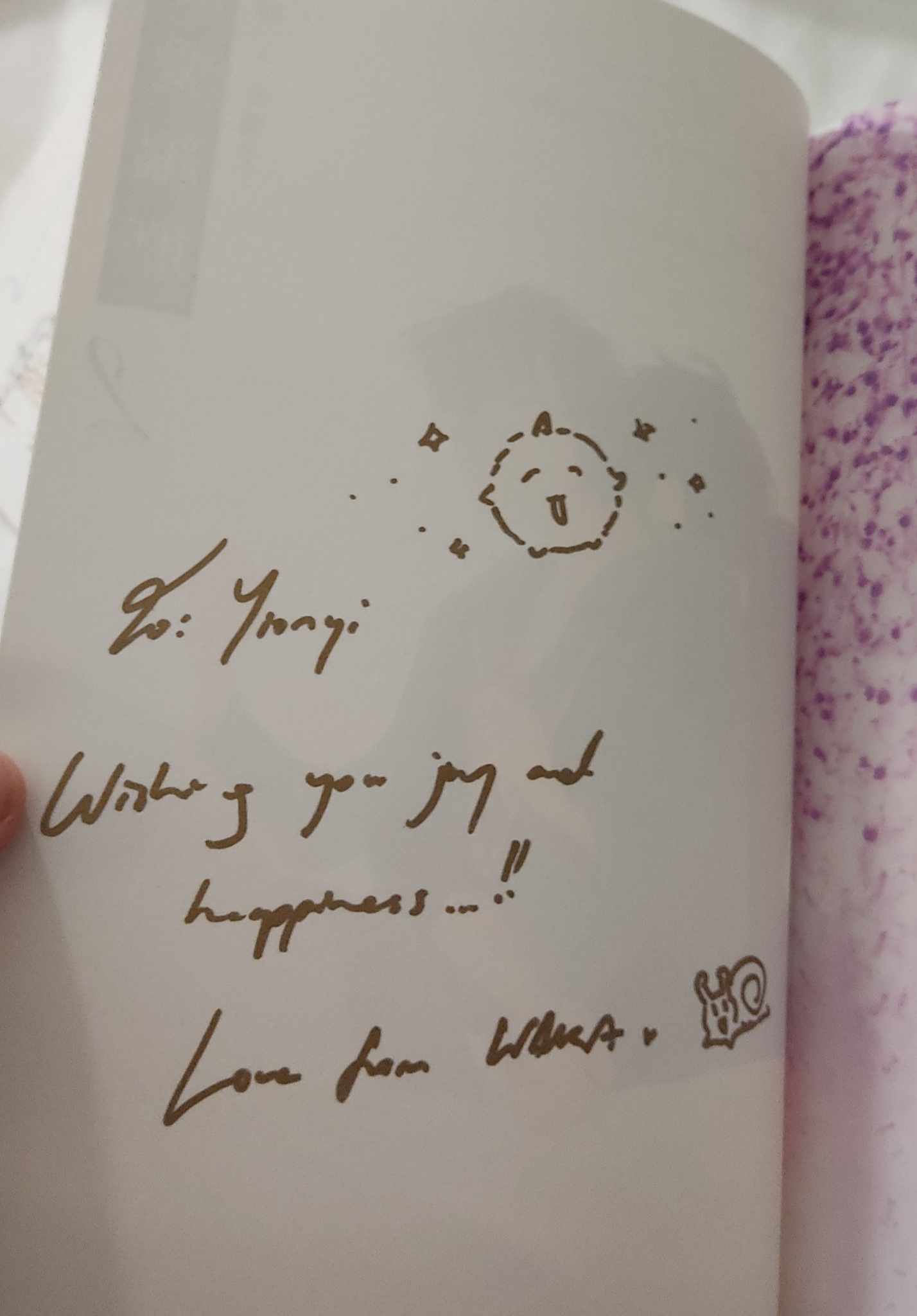 closeup shot of a book's inside cover page with a handwritten message. the message is written in gold ink and says "to: ynagi, wishing you joy and happiness... love from whale." there's a small, cute doodle of a creature and a whale next to the message. the background shows a slightly textured paper.