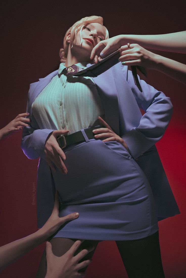 a woman with blonde hair in business attire (light purple jacket, skirt and shirt with a tie) is posing. several disembodied hands are touching or holding her clothing. the background is a dark red color.