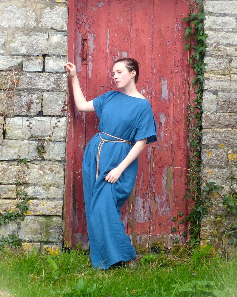 a woman stands against a weathered red wooden door set into a stone wall. she is wearing a long, loosefitting,  blue tunicstyle dress that resembles ancient greek clothing. the dress is cinched at the waist with a simple cord.  the scene is outdoors, with grass and ivy growing around the stonework. the overall atmosphere is rustic and evocative of a historical or period setting. the woman's pose is relaxed yet graceful.