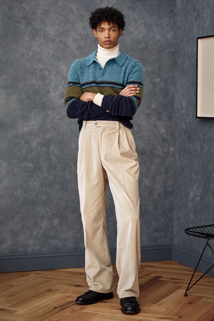 a fullbody shot of a male model showcasing a casual autumnal outfit. he is wearing a teal and olive striped poloneck sweater over a white turtleneck, paired with beige corduroy pleated trousers and black shoes. the model stands against a textured dark gray background, with a wooden floor visible. a simple black metal chair is partially visible in the background. the overall aesthetic is one of relaxed yet stylish autumnal fashion.