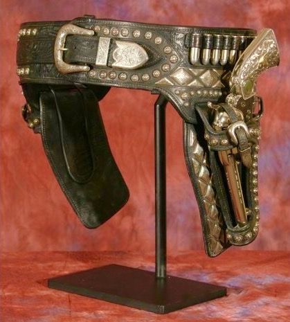 a detailed closeup shot of an antique, ornate gun belt with two holsters and pistols. the belt is made of leather, heavily studded with silver, and features engraved details.  the pistols appear to be small, possibly .22 caliber. the entire ensemble is displayed on a black stand against a red backdrop. the image highlights the craftsmanship and intricate details of the piece.