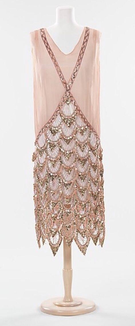 a vintage 1920s flapper dress on a mannequin. the dress is made of a light pink silk and is heavily beaded with sequins and gold thread. the dress has a loose, flowing silhouette and a dropped waistline. it is sleeveless and has a deep vneckline. the dress is a beautiful example of the flapper style, which was popular in the 1920s. the dress is on display in a museum.