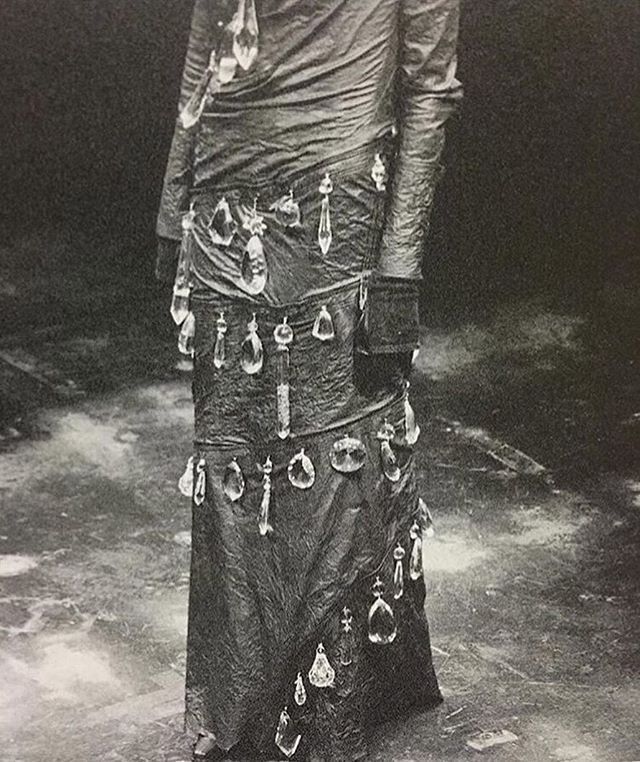 a black and white photo shows a person wearing a draped gown adorned with crystal ornaments. the dress has long sleeves and a floorlength skirt, with the crystals attached in rows.