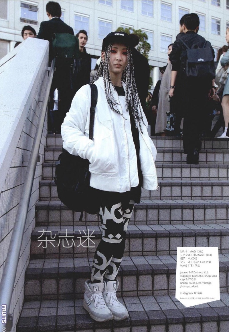 a young adult with long white braids and a black baseball cap poses for the camera. they are wearing a white bomber jacket, black printed leggings, and white sneakers. they have a black backpack slung over one shoulder. they are standing on a set of stairs in an urban setting. there are other people walking in the background. the image is captioned with japanese characters and english text.