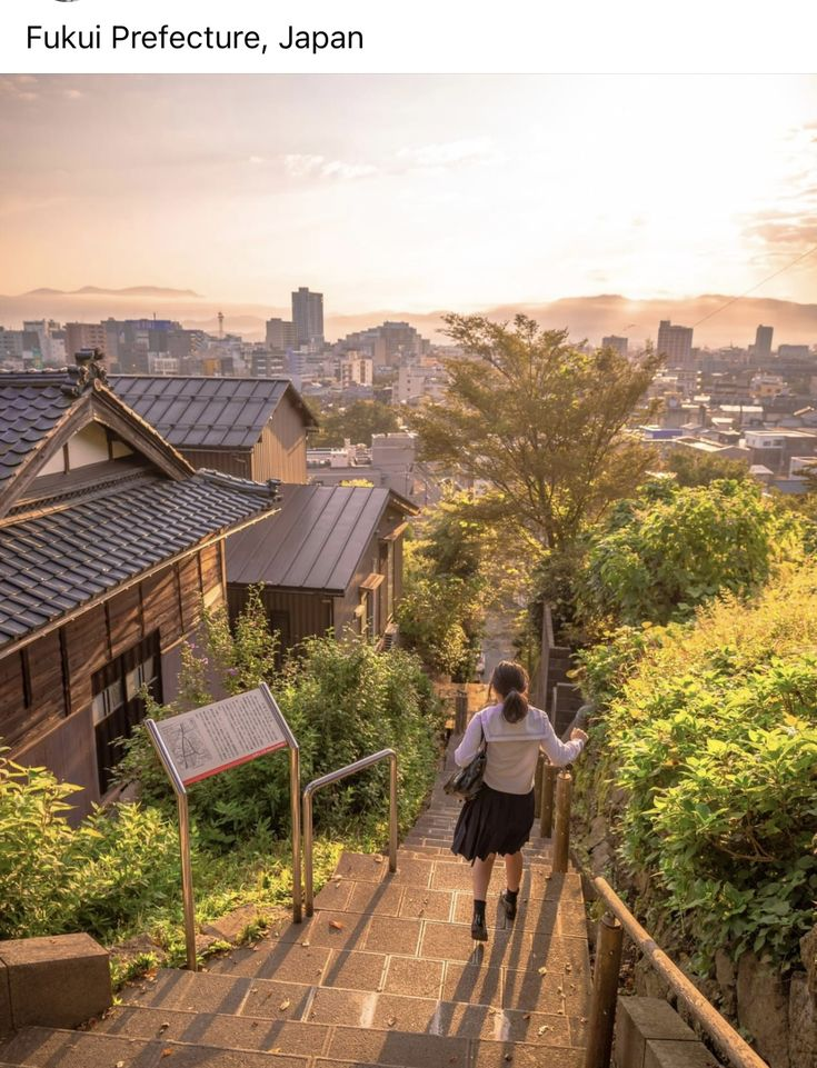 a medium shot of fukui prefecture, japan, showing a cityscape at sunset. a person in a school uniform walks down a set of stairs with railings. buildings and trees are seen in the background.