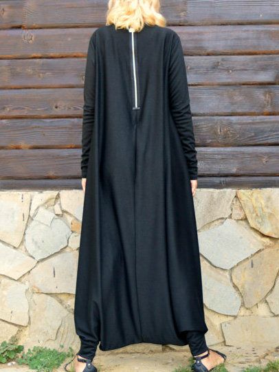 a woman from the back wearing a long, black jumpsuit with long sleeves and a zipper down the back. she is standing outside in front of a wall made of stacked stones and wearing black sandals.