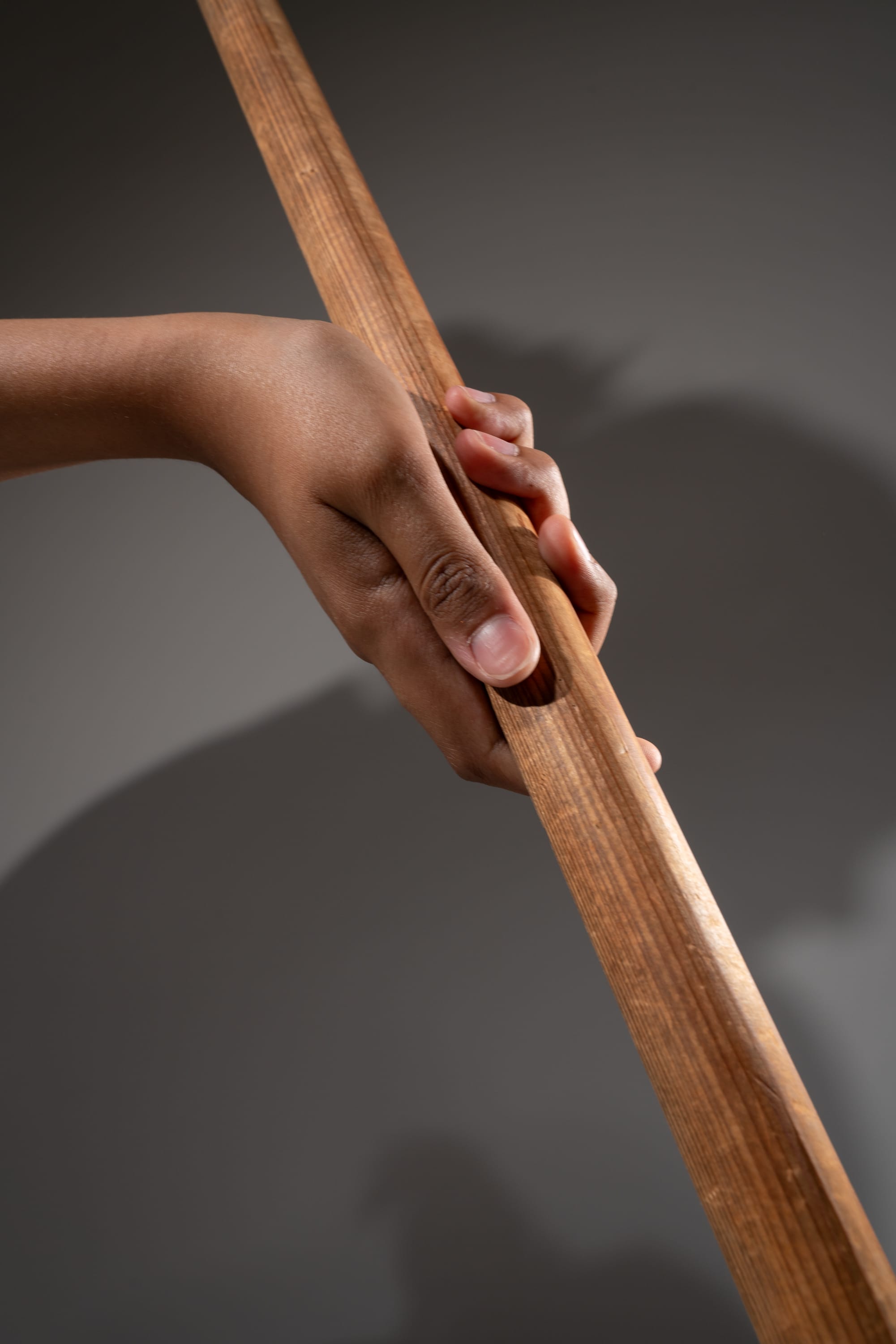 a closeup view of a hand tightly gripping a wooden stick. the stick is smooth and polished. the background is a neutral gray color, and the lighting is soft and diffused. there is a shadow of the stick and hand cast on the background.