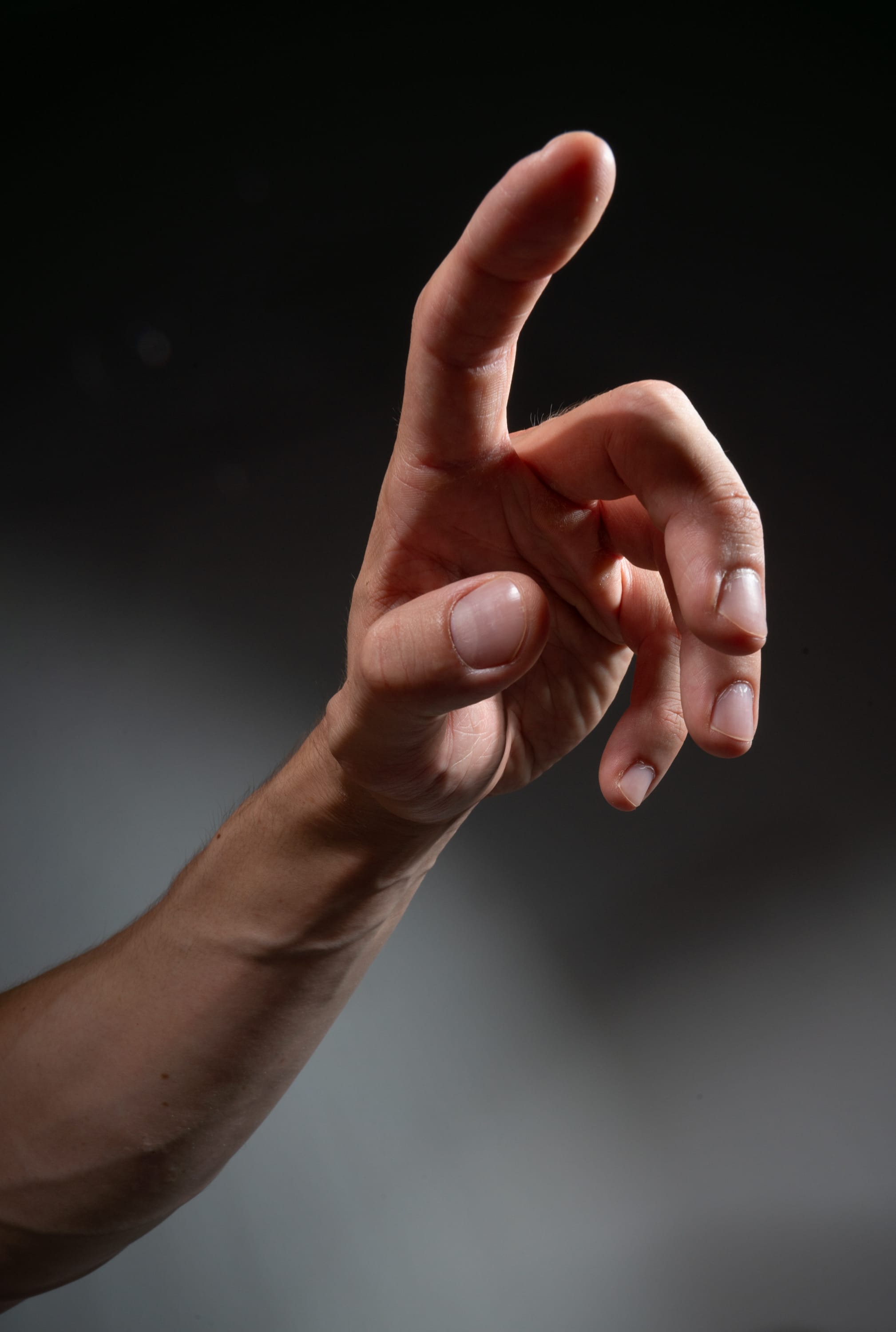 the image is a closeup shot of a man's hand pointing upwards against a dark gray background. the hand is illuminated with studio lighting, highlighting the details of the skin and veins. the image focuses on the gesture of pointing, suggesting direction, communication, or emphasis.