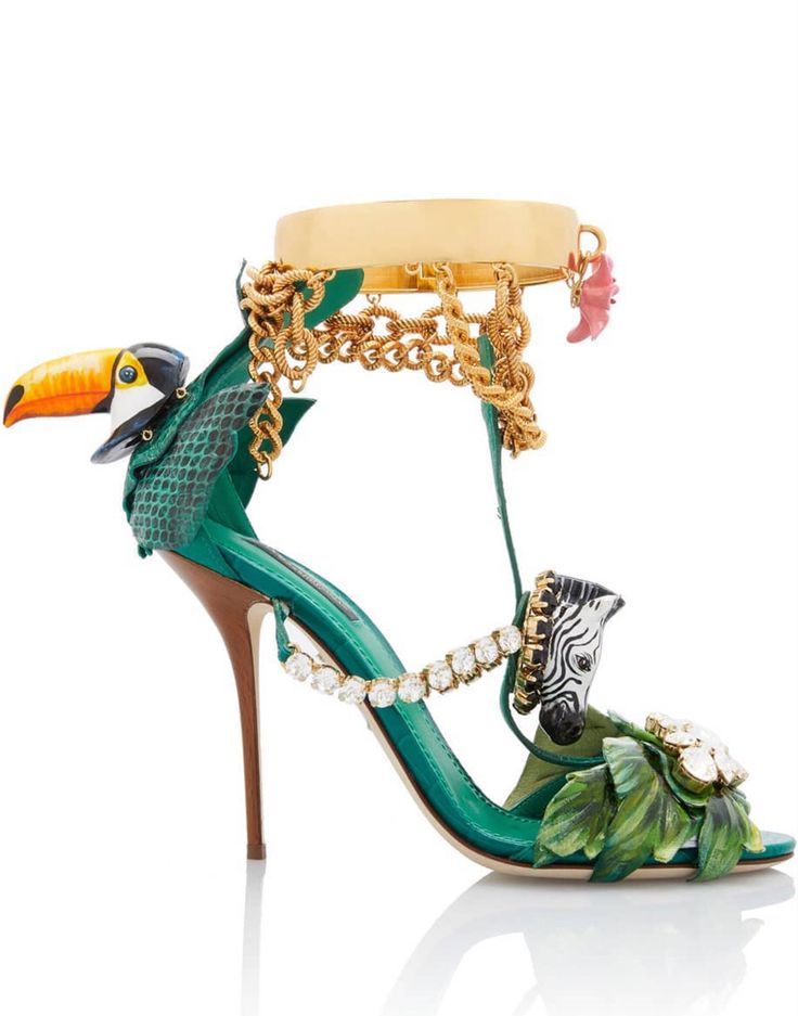 the image showcases a pair of striking dolce & gabbana highheeled sandals.  the shoes are a vibrant green, adorned with an array of exotic embellishments.  these include a toucan's head at the ankle, a zebra head on the strap, lush green leaf appliqués, and a substantial gold chain and cuff detail.  the heels are also decorated with clear crystals and rhinestones, adding to the overall extravagant and eyecatching design. the sandals are positioned on a reflective white surface, highlighting their details and craftsmanship.  the style is distinctly bold and luxurious, reflecting the brand's signature aesthetic.