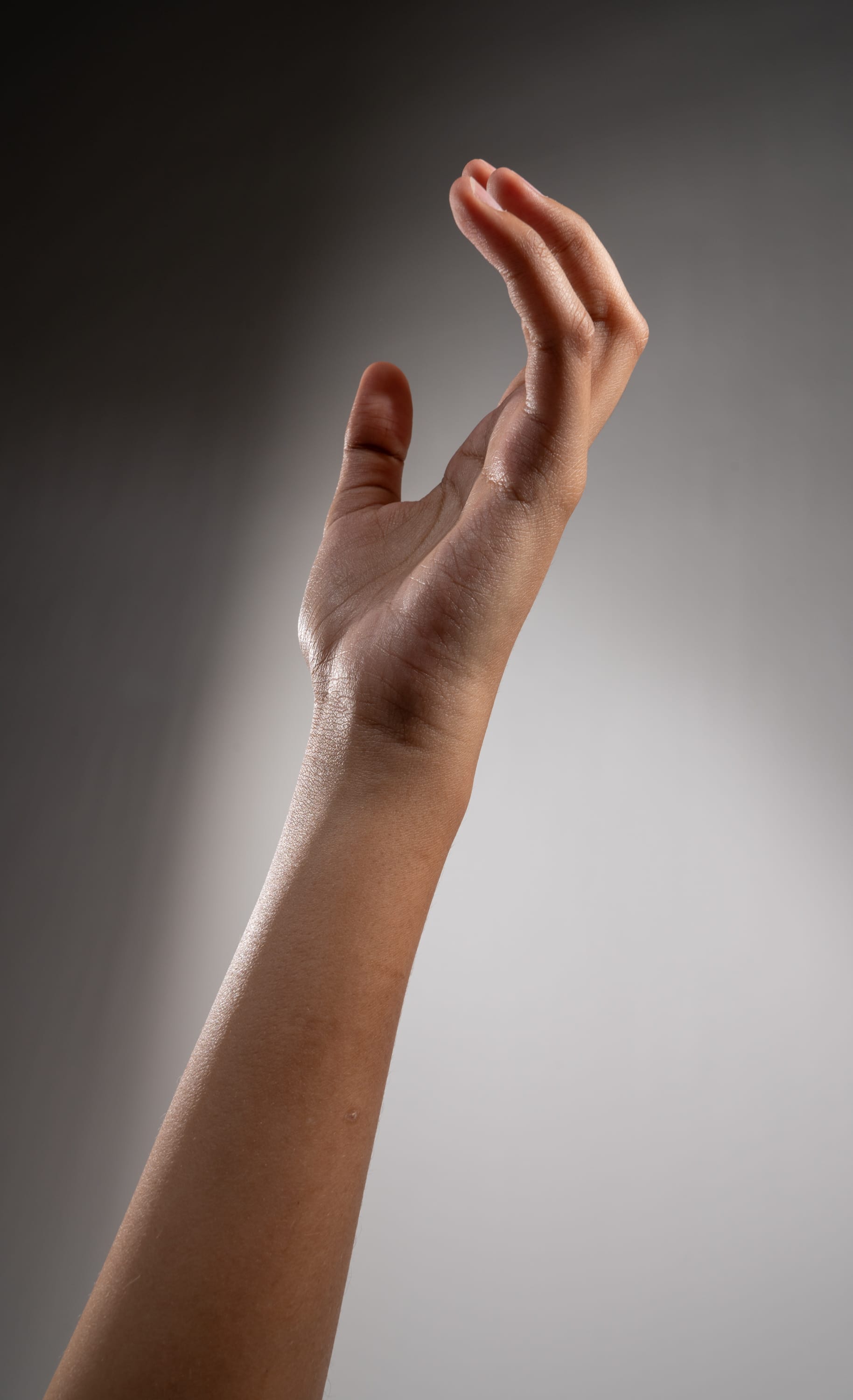 a left hand and arm extended up against a gray background. the arm is outstretched with the palm facing the viewer and fingers curled inward. the hand is lit with soft studio lighting.