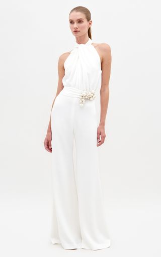 a woman is standing in a white, halter jumpsuit in a studio setting. she is looking straight ahead. the jumpsuit is a simple yet elegant style.  her hair is pulled back, revealing her beautiful features. she has a simple, elegant look.