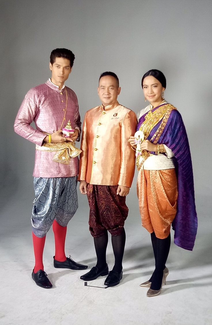 the image shows three people dressed in traditional thai clothing. the woman is on the right wearing a purple and orange outfit with a white sash. the man on the left is wearing pink and silver with a sash. the man in the middle is wearing orange and brown, with a sash. they are all standing in front of a white background.