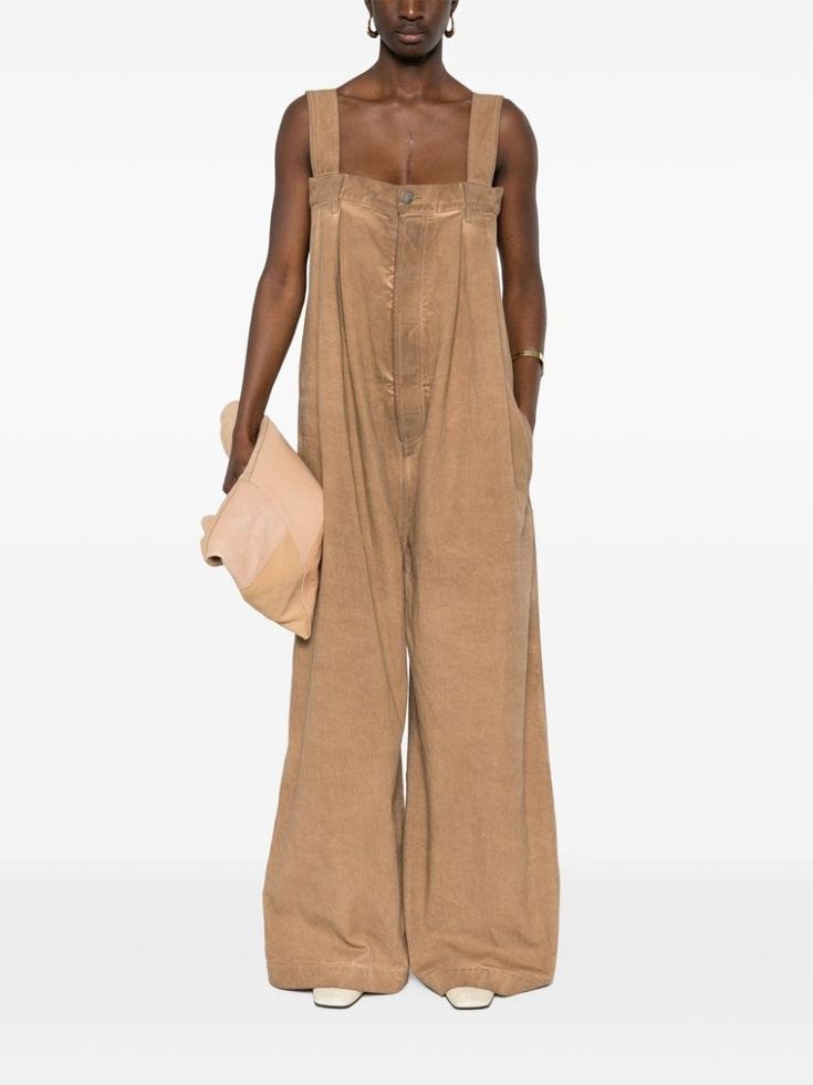 a woman wearing a tan wideleg jumpsuit. she is holding a lighttan bag and wearing white shoes. she is standing in front of a white background.